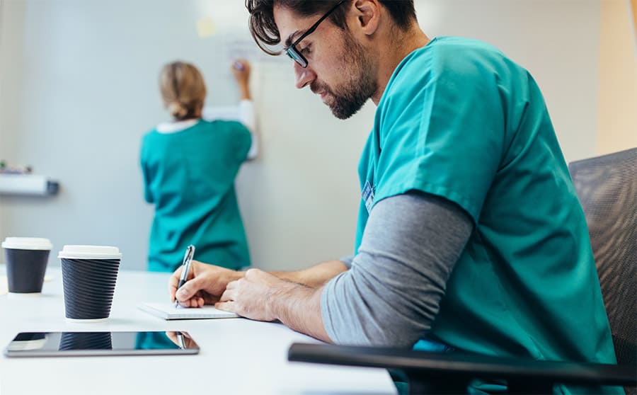 A nurse sitting at a desk writing notes, while another writes on a board in the background